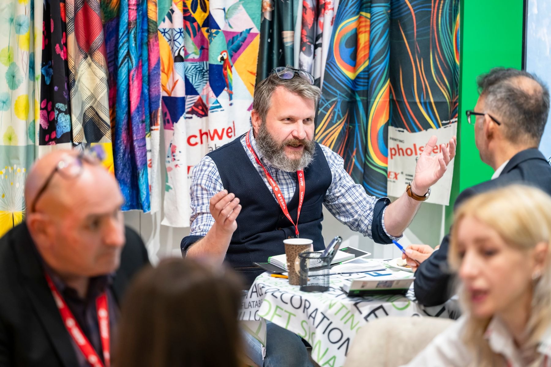 Exhibitor in conversation with visitors at a stand surrounded by printed textile samples