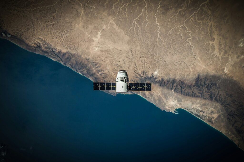 Spacecraft with deployed solar arrays in low Earth orbit above a desert coastline, an example of satellite design using advanced composite materials