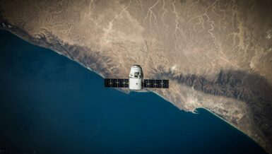 Spacecraft with deployed solar arrays in low Earth orbit above a desert coastline, an example of satellite design using advanced composite materials